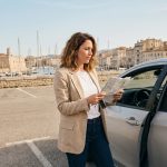 Femme debout à côté d'une voiture de location argentée près du Vieux-Port de Marseille, tenant une carte routière et des clés de voiture