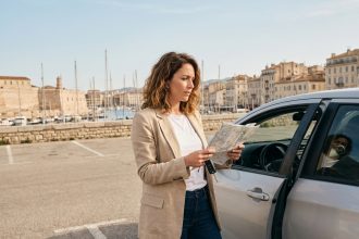 Femme debout à côté d'une voiture de location argentée près du Vieux-Port de Marseille, tenant une carte routière et des clés de voiture