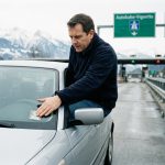 Conducteur collant une vignette autoroutière suisse sur le pare-brise d'une voiture au bord d'une autoroute alpine